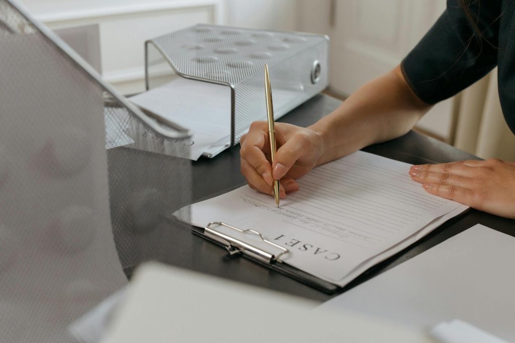 Close-up of a woman writing on documents with a gold pen in an office environment.