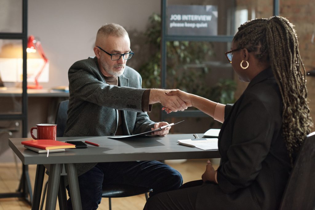 Mature manager shaking hands with young woman while they sitting at table during job interview